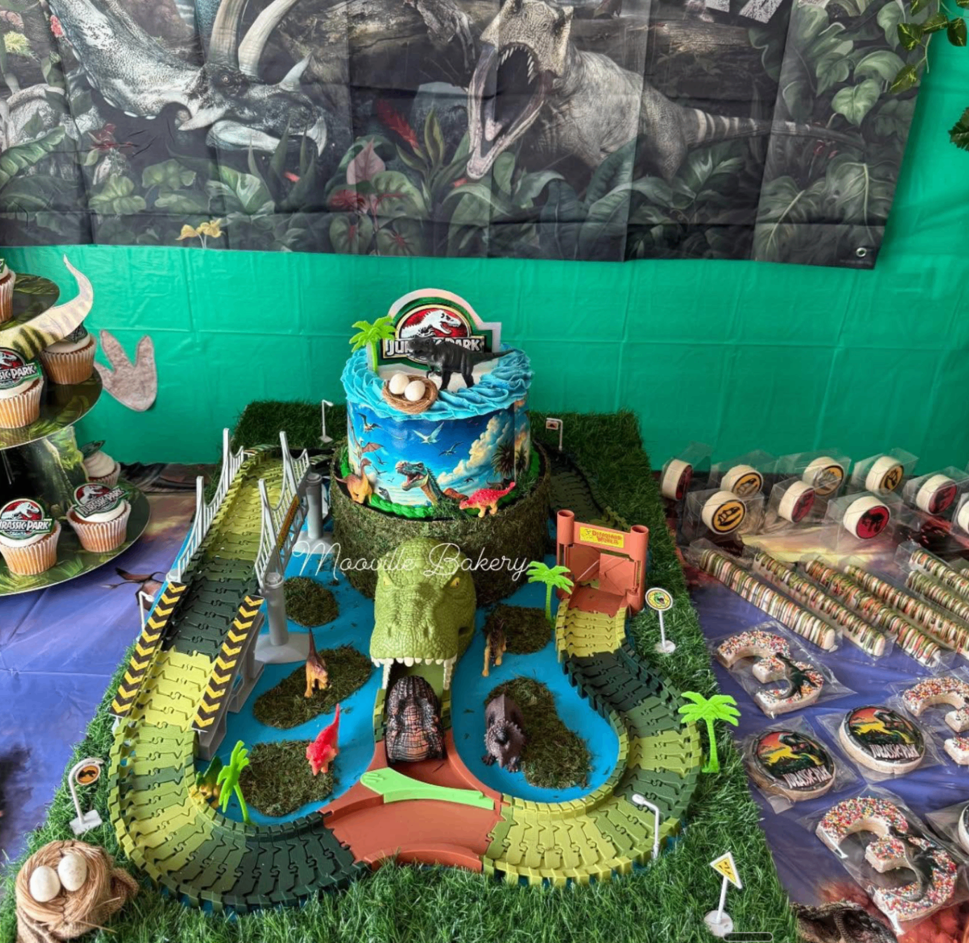 Mooville Bakery Jurassic Park dessert table with themed cake, cupcakes, cookies, and dinosaur scene.